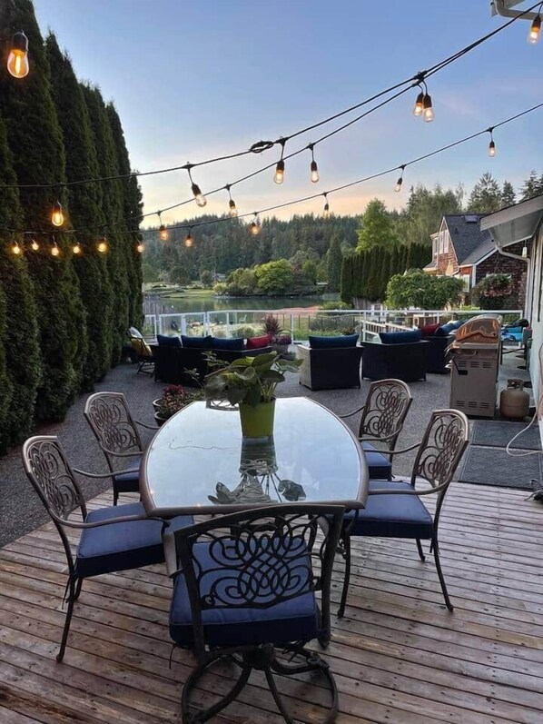 A wooden deck with a glass-top table surrounded by chairs, string lights overhead, and a view of a lake and trees in the background.