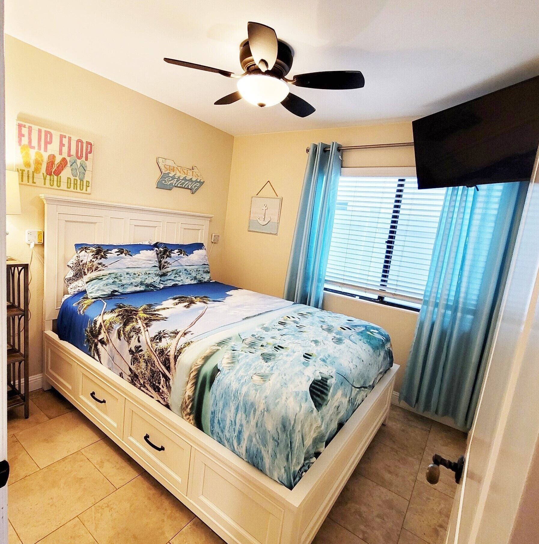 Un dormitorio con cama, ventilador de techo, ventana con cortinas y televisor de pared.