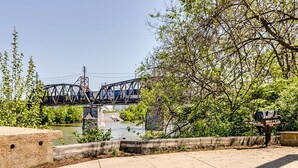 A riverside walkway with a stone fire pit, a bridge in the background, and lush greenery. - Stunning River View Condo Near Downtown With Free Covered Parking (Nashville)