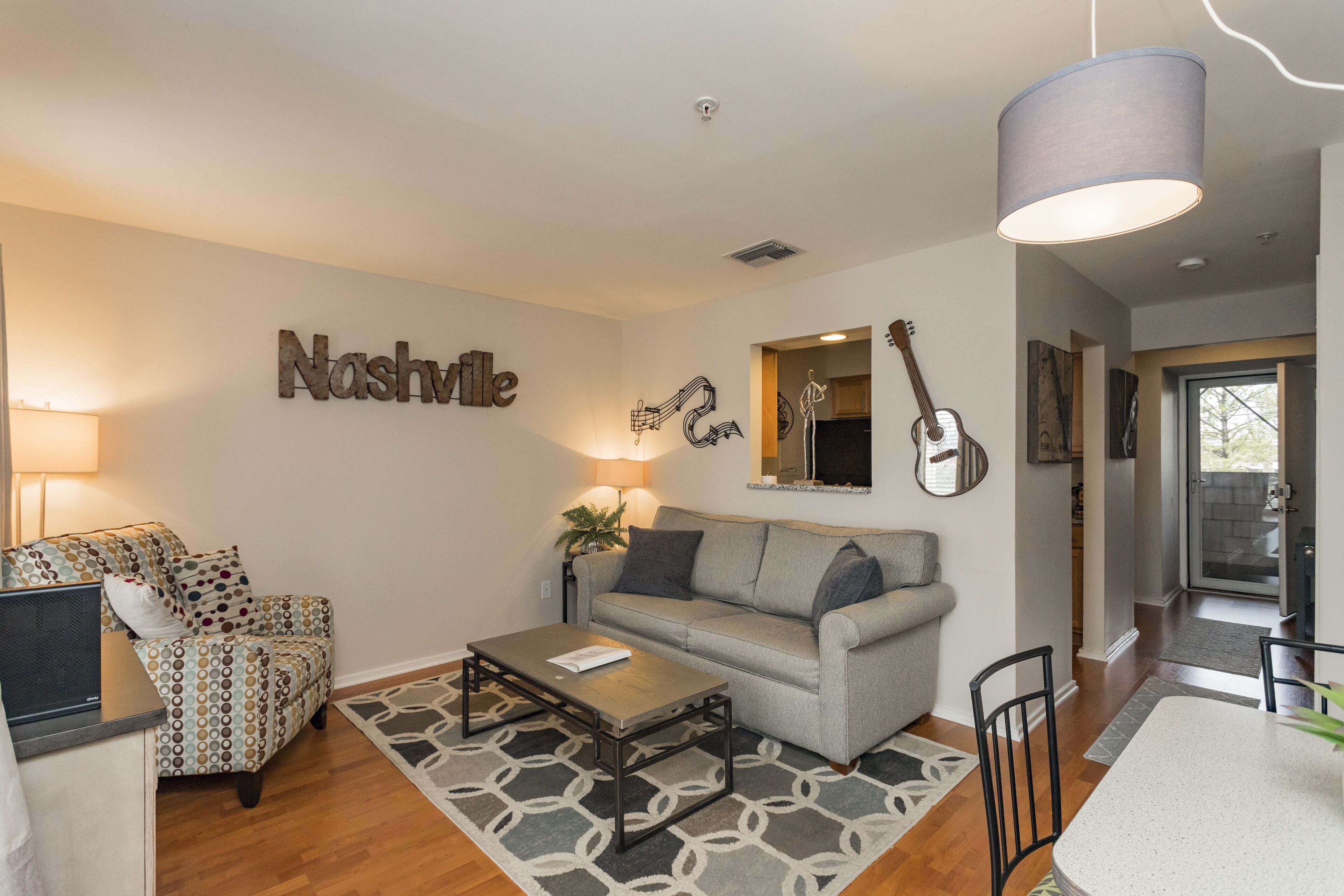 A living room with a grey sofa, a patterned armchair, a coffee table, and a dining table with chairs. There is a guitar on the wall and a lamp above the sofa.