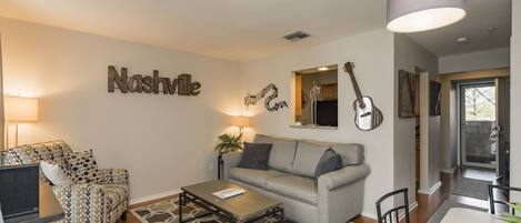 A living room with a grey sofa, a patterned armchair, a coffee table, and a dining table with chairs. There is a guitar on the wall and a lamp above the sofa.