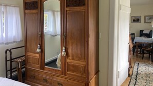 A traditional wooden armoire with a large mirror, a small table with a chair, and a patterned rug in a room with a door leading to another area. - Private Garden Cottage In Mendocino's Historic Preservation District (Mendocino)