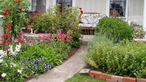 A garden with a variety of flowers and plants, a pathway, and a wicker sofa on the porch. - Private Garden Cottage In Mendocino's Historic Preservation District (Mendocino)