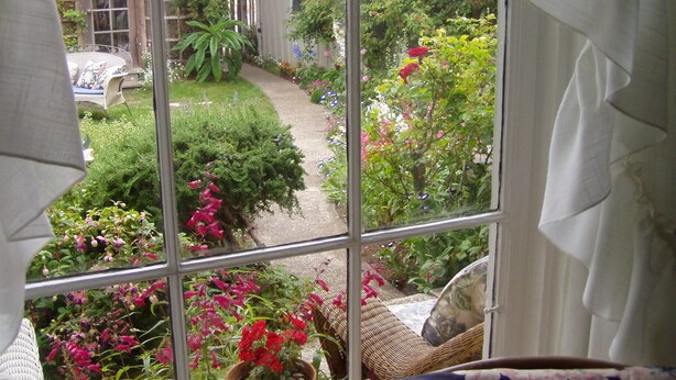 A view through a window of a garden with a pathway, various plants, and a wicker chair. - Private Garden Cottage In Mendocino's Historic Preservation District (Mendocino)