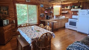 Interior de una cabaña de madera con una mesa de comedor, sillas, una zona de cocina con estufa, refrigerador y una ventana con persianas.