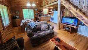 Interior de una cabaña con comedor de madera, un sofá, un televisor y una escalera.