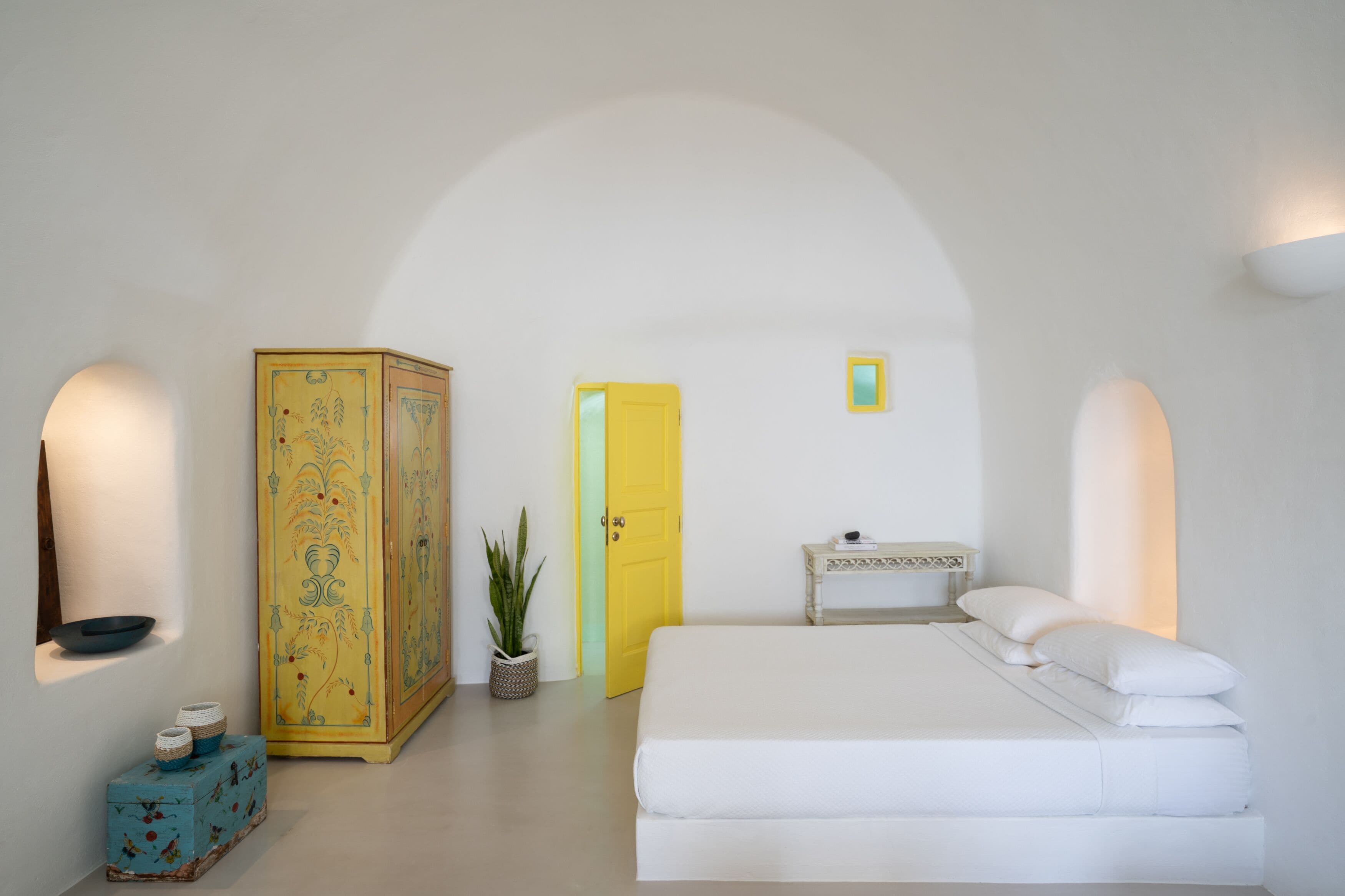 A minimalist room with a bed, a yellow door, a small table, and a decorative cabinet.
