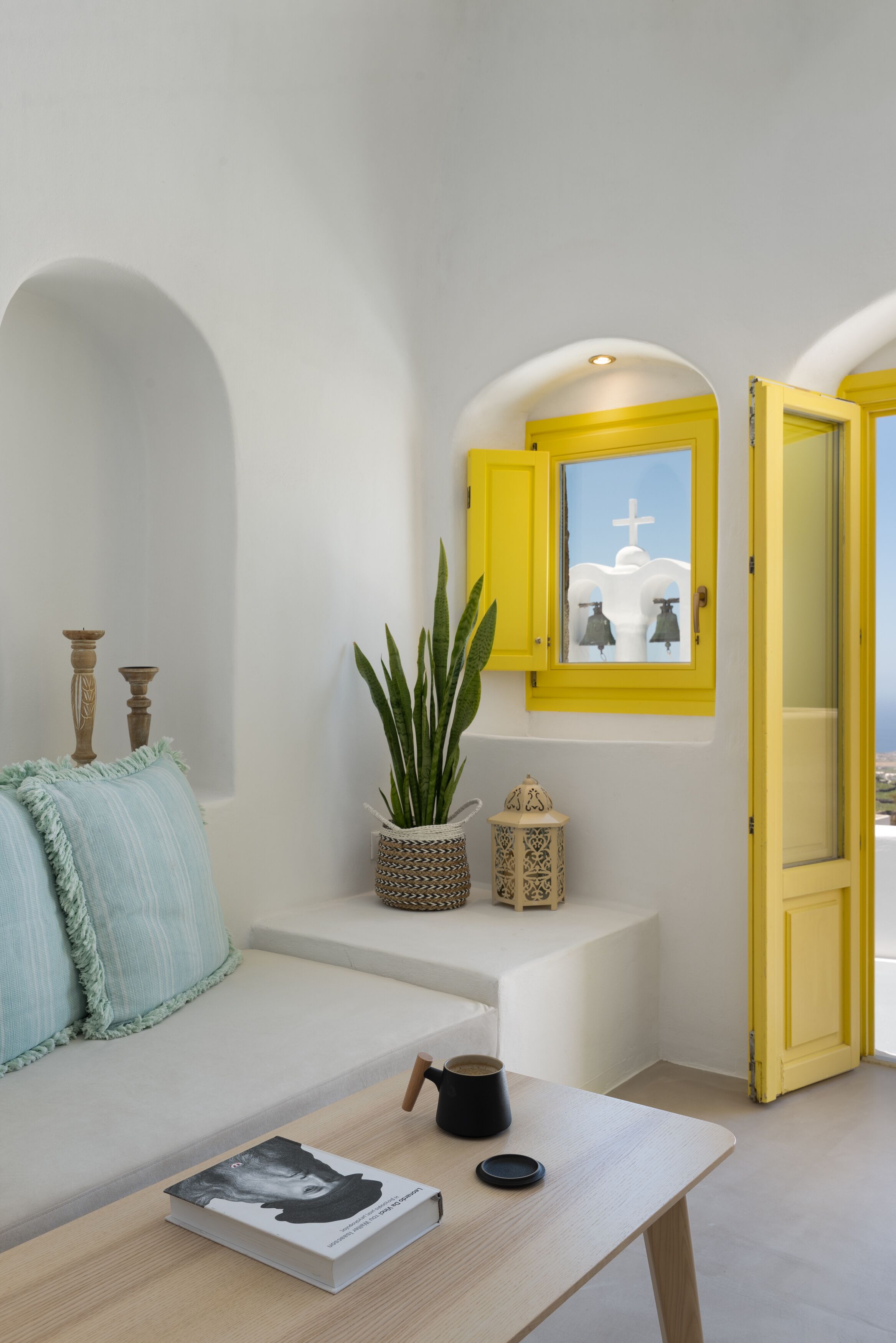 A room with a white sofa, a small table with a book and a cup, a potted plant, and a yellow-framed window with a cross and bells.