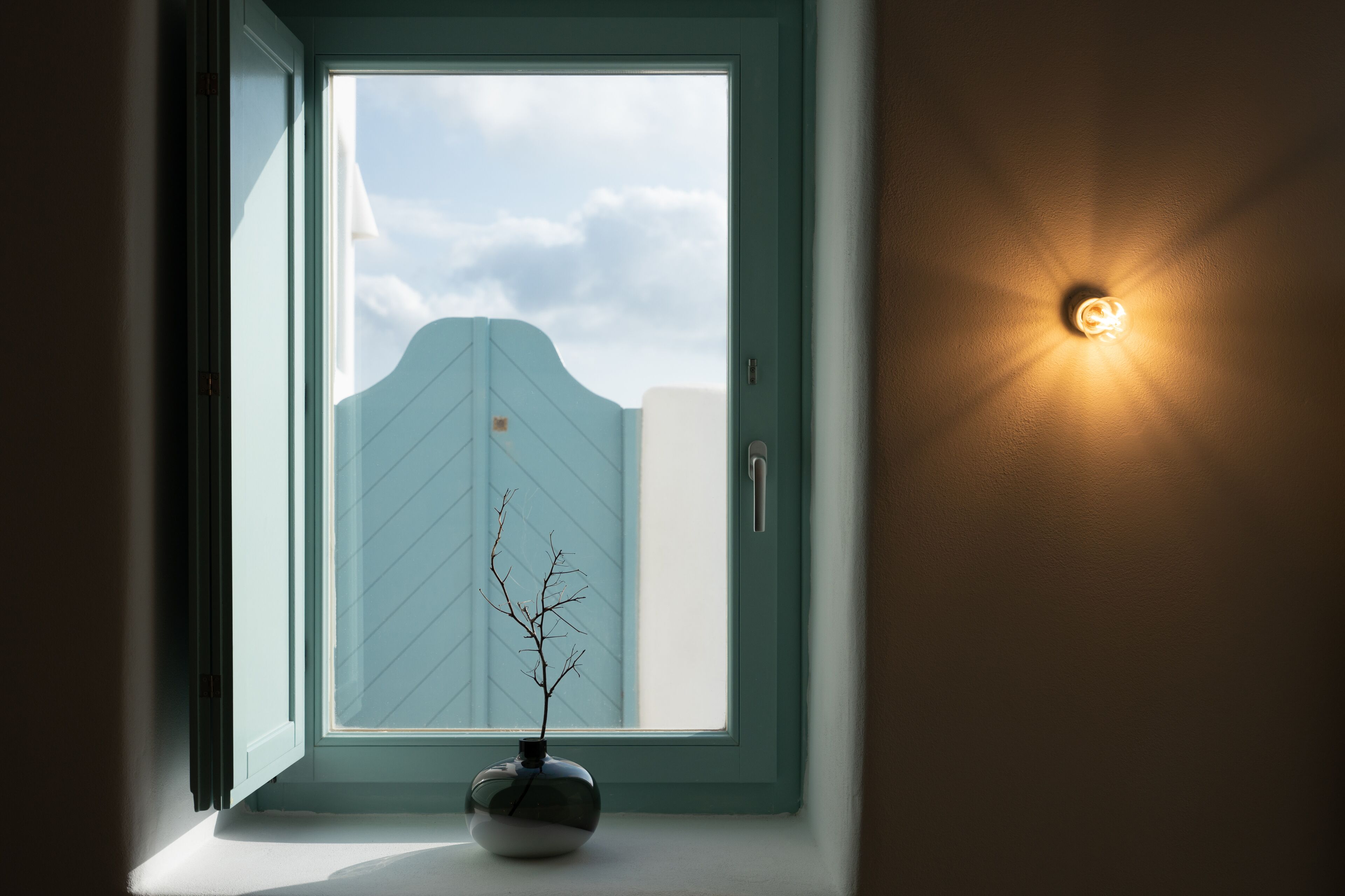 A room with a window, a wall-mounted light, and a vase with a branch.