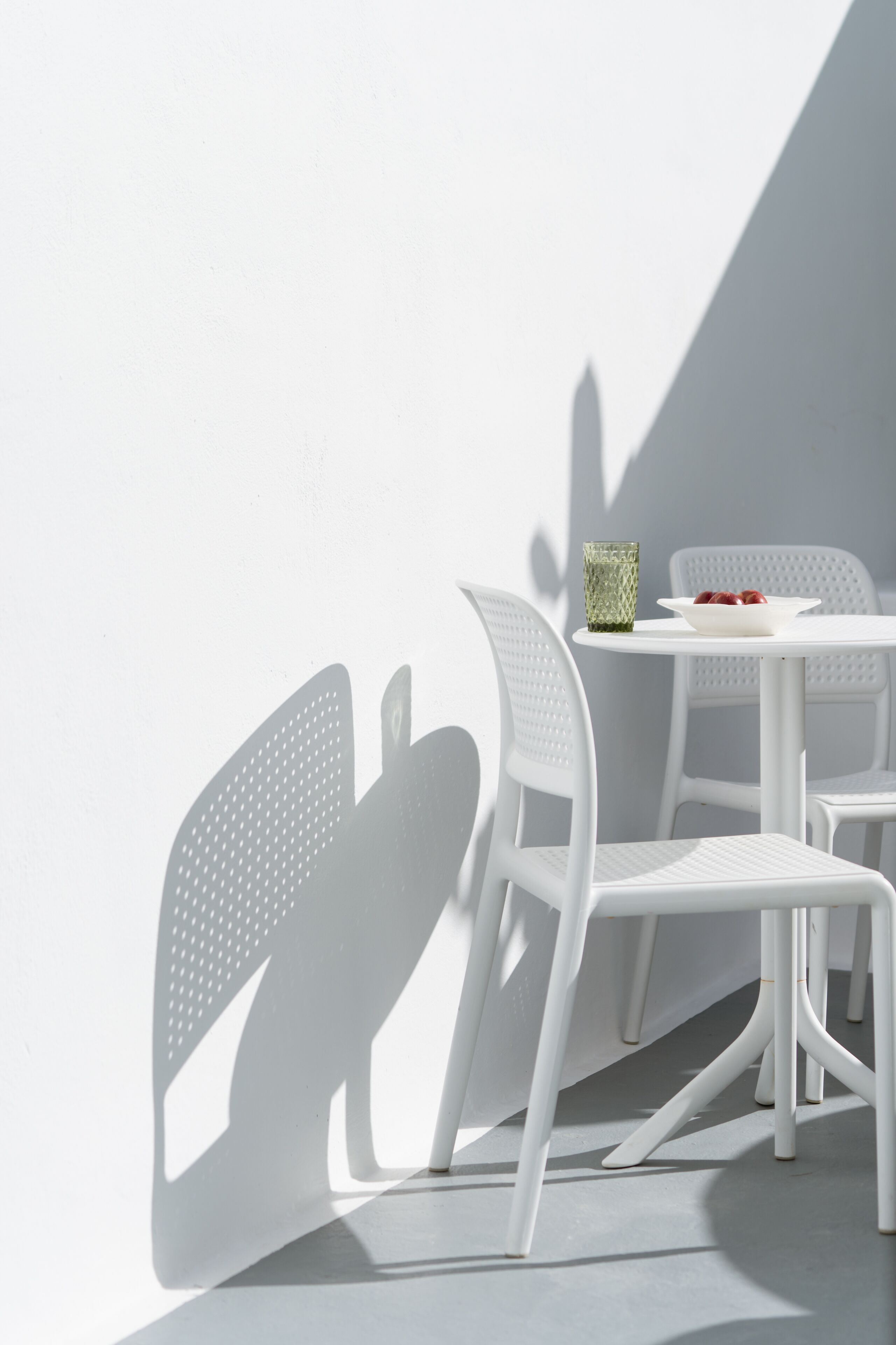 Uma área de estar externa com mesa e cadeiras brancas, um recipiente de vidro e uma sombra projetada na parede.