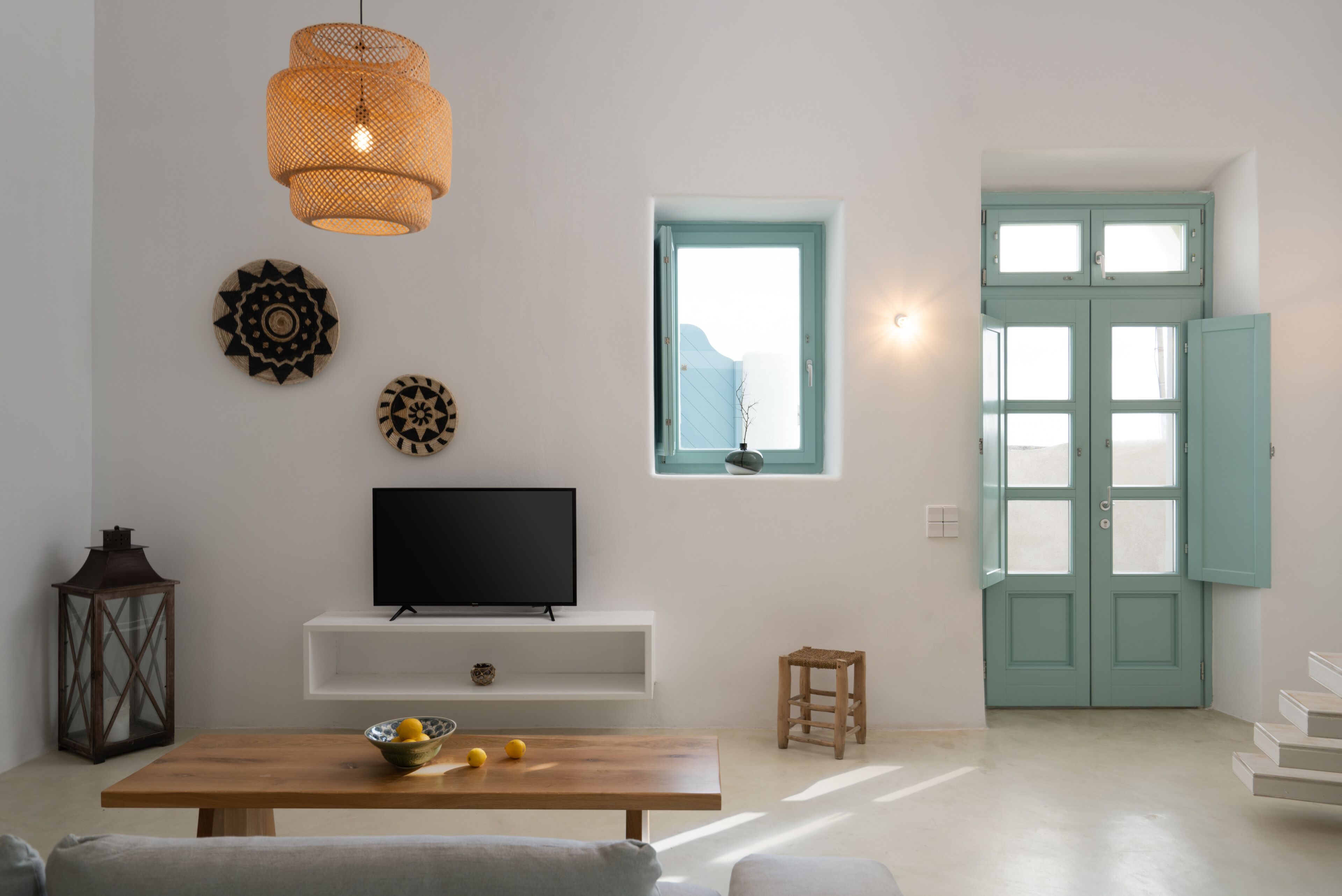 A modern living room with a wooden table, a TV, decorative wall art, a wicker pendant light, a small stool, and a teal door.