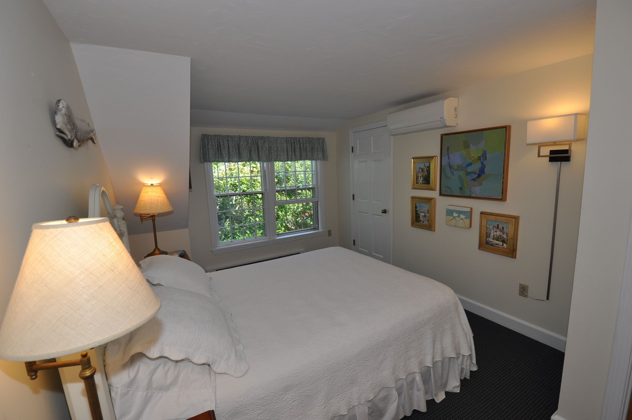 A neatly arranged bedroom with a bed, a window with a view of greenery, a lamp, and wall-mounted pictures.