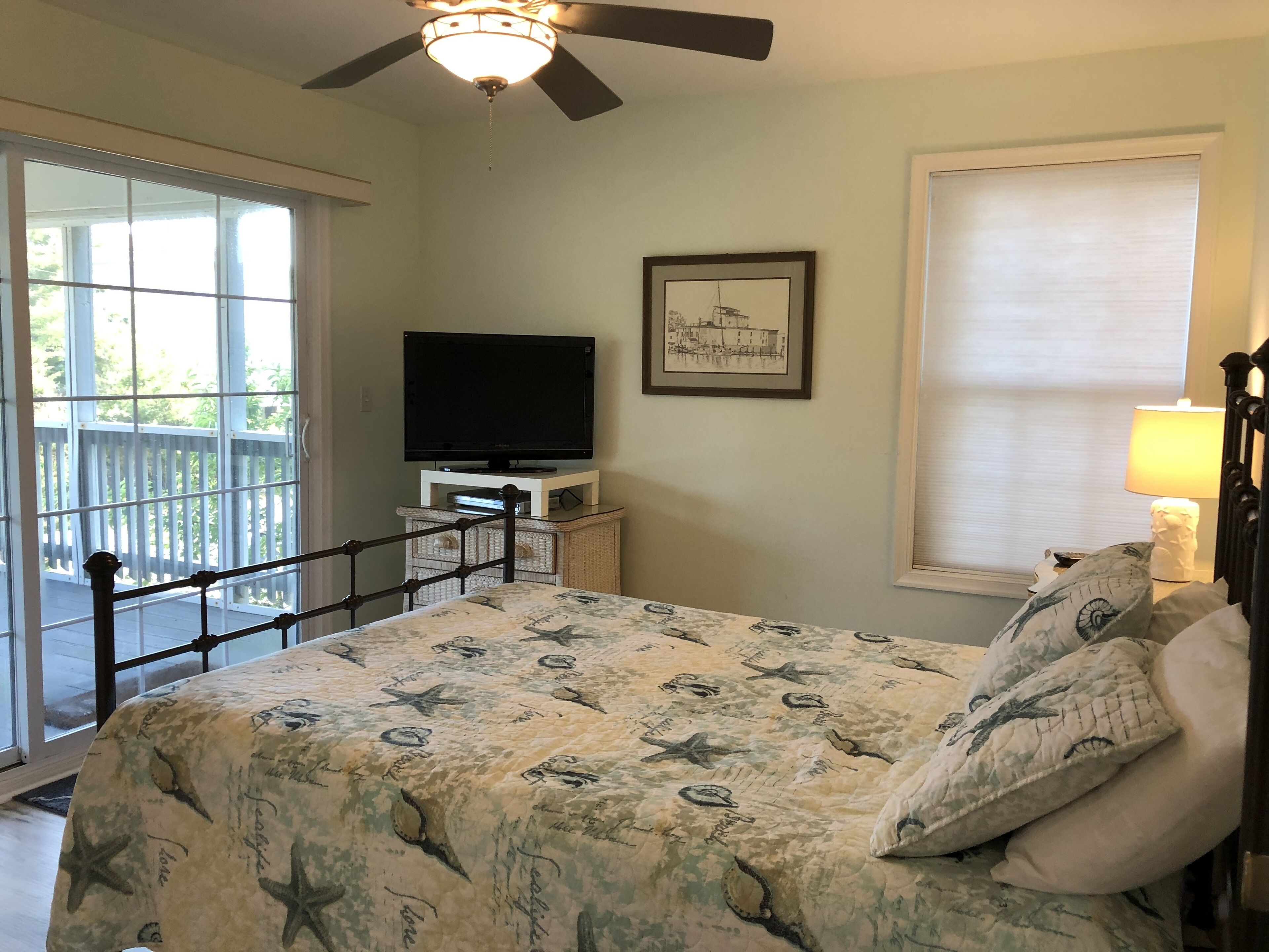 A bedroom with a bed, a television on a stand, a framed picture on the wall, a window with a blind, and a ceiling fan.