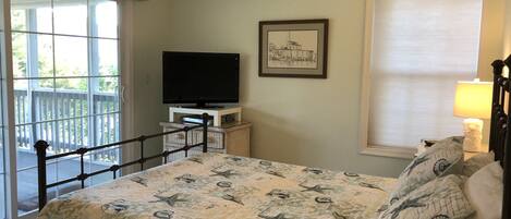A bedroom with a bed, a television on a stand, a framed picture on the wall, a window with a blind, and a ceiling fan.