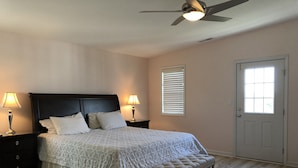 Un dormitorio con una cama, dos mesitas de noche con lámparas, un ventilador de techo, una ventana con persianas y una puerta.