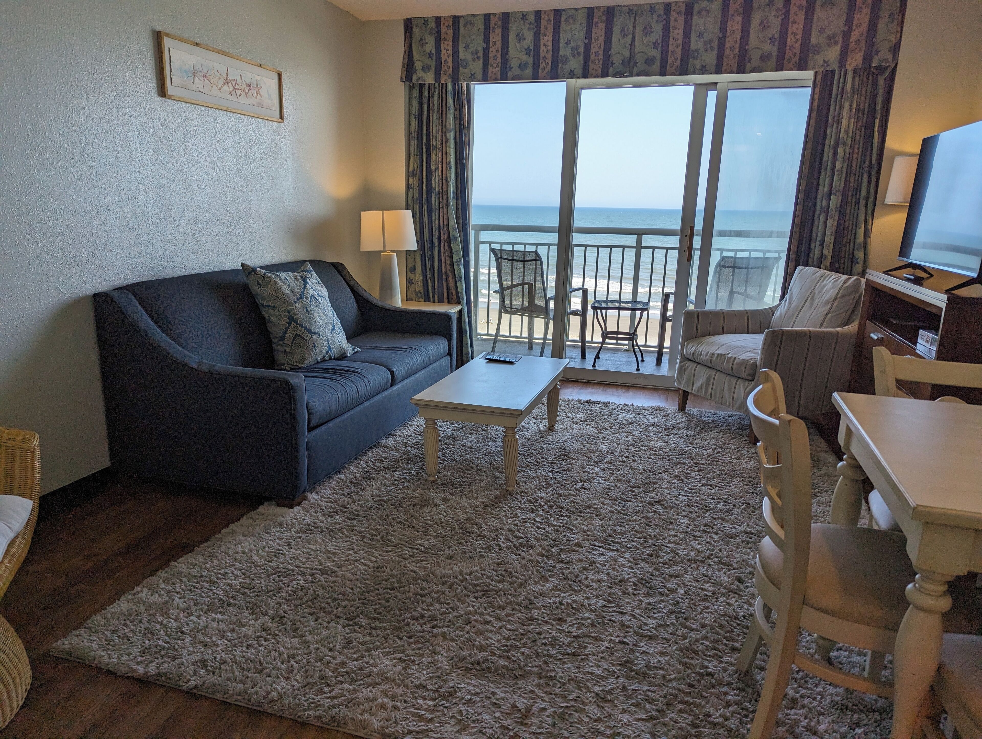 A living room with a sofa, a coffee table, and a TV. There is a balcony with a view of the ocean.