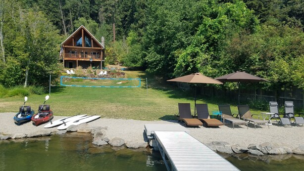 Une cabane avec une terrasse donnant sur un lac, des kayaks, des chaises longues et des parasols.