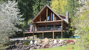 Une cabane en rondins dotée d’un balcon, entourée d’un jardin aménagé avec des rochers et un drapeau.