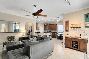 A spacious living room with a grey sectional sofa, a dark ottoman, and a ceiling fan. There is a kitchen area with wooden cabinets and a wine rack.