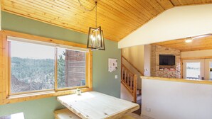 A wooden interior with a dining table and bench, a window with a view of a forest, and a stone wall with a flat-screen TV. - Pikes Peak Ranch - Elk Ridge Cabin (Woodland Park)