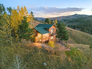 Une cabane en rondins  à deux étages, dotée d’un toit végétalisé, entourée d’arbres et de collines.