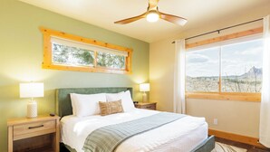 A bedroom with a bed, two bedside tables, a ceiling fan, a window with a view of a mountainous landscape, and a wooden floor. - Pikes Peak Ranch - Elk Ridge Cabin (Woodland Park)