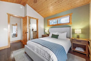 A bedroom with a wooden ceiling, a large bed with a white and blue patterned bedspread, a nightstand with a lamp, and a window with a view of trees. - Pikes Peak Ranch - Elk Ridge Cabin (Woodland Park)
