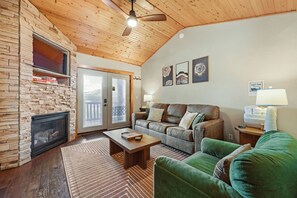 A cozy living room with a stone fireplace, a leather sofa, a green armchair, and a ceiling fan. - Pikes Peak Ranch - Elk Ridge Cabin (Woodland Park)