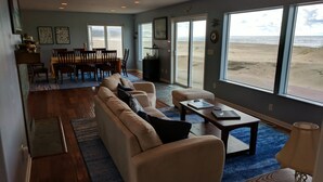 Een ruime woonkamer met een grote bank, een eethoek met een tafel en stoelen, en grote ramen met uitzicht op het strand.
