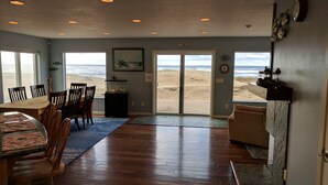 Een ruime kamer met houten vloer, een eettafel met stoelen, een groot raam met uitzicht op een strand en een deur die naar het strand leidt.