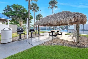 An outdoor patio with a thatched-roof structure, a barbecue grill, a picnic table, and a trash can. - Gorgeous Waterview, Walking Distance to Restaurants, Shops & More!! (Marco Island)