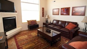 A living room with a fireplace, a large window, a brown leather sofa, a coffee table, and framed artwork on the wall.