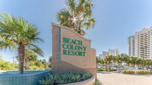 Une enseigne indiquant l’emplacement du Beach Colony Resort, offrant une vue dégagée sur les environs, notamment sur une piscine et des palmiers.