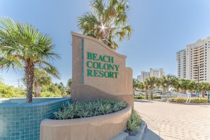 Beach Colony Resort -hotellin kyltti, josta avautuu selkeä näkymä ympäröivään alueeseen, mukaan lukien uima-allas ja palmuja.