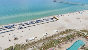 Eine Vogelperspektive zeigt einen Strand mit einer langen Mole, die ins Meer führt, einen sandigen Strandabschnitt mit Reihen blauer Sonnenschirme und Liegestühlen sowie ein Schwimmbecken mit Menschen, die das Wasser genießen.
