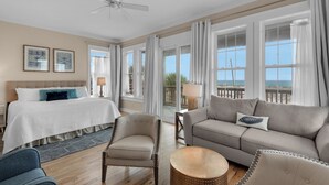 A hotel room with a bed, a sofa, two armchairs, a round coffee table, and a balcony with a view of the beach.