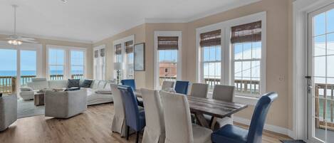 A spacious living room with a large dining table, comfortable seating, and large windows offering a view of the ocean.