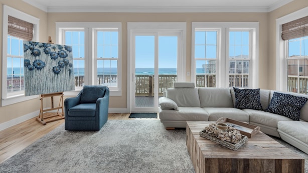 A spacious living room with a large window overlooking the beach, a blue armchair, a beige sofa, and a wooden coffee table.