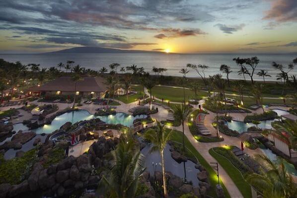 A resort with multiple swimming pools, palm trees, and a view of the ocean at sunset.