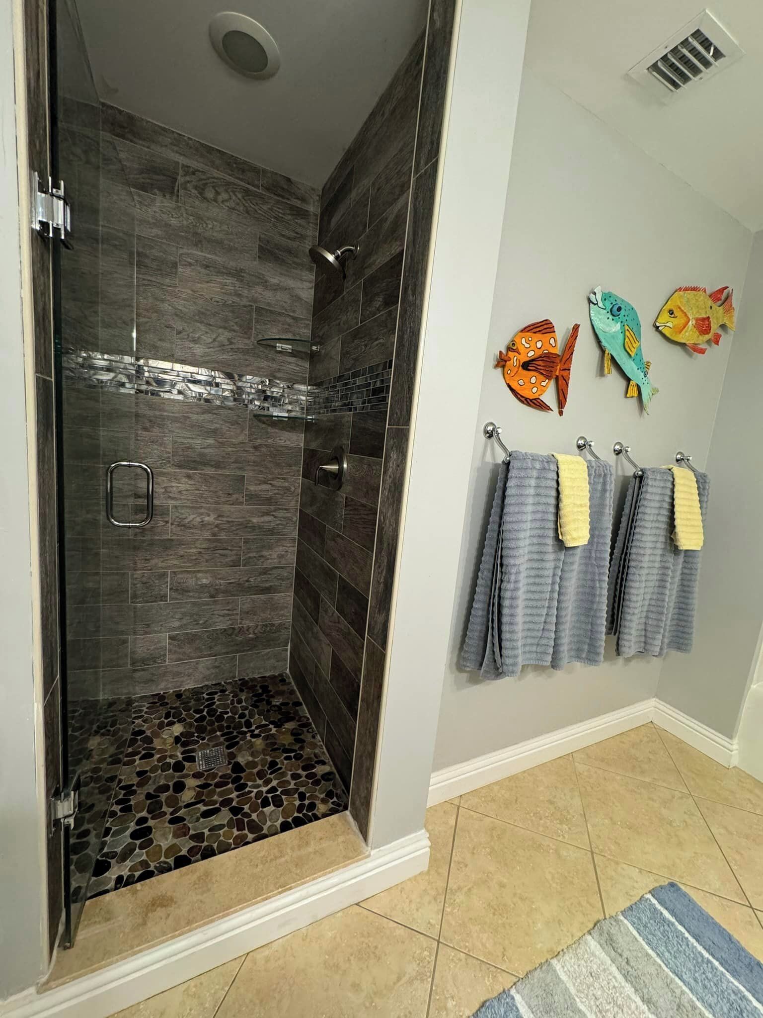 Um chuveiro sem barreiras com azulejos escuros e piso em mosaico, um porta-toalhas com várias toalhas, e uma decoração de peixe na parede.