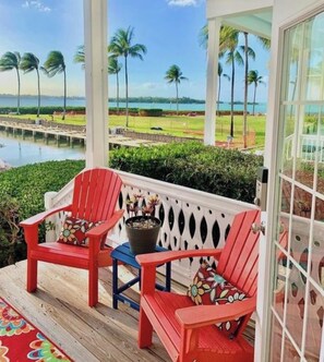 A porch with two red Adirondack chairs and a blue chair, a potted plant, and a view of a golf course and water. - SPECTACULAR Indigo Reef Waterfront Home #41 - Marathon, Florida (Marathon)