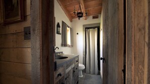 Une salle de bain rustique avec des murs en bois, un sol en pierre, un meuble-lavabo en bois, un évier, des toilettes et un espace douche.