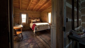 Une chambre confortable dans un chalet en rondins, avec un plafond en bois, un lit recouvert d’une couverture rouge, un bureau avec une chaise et une petite table sur laquelle sont posés des livres.