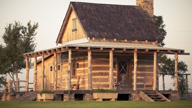Une cabane rustique en rondins de bois, dotée d’un porche, d’une cheminée et d’une petite fenêtre.