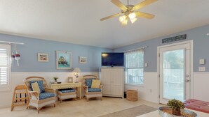A room with a ceiling fan, a television, wicker furniture, a small table, and a door with a window. - MERMAID - Largest Heated Pool, Walk to Private Beach, GolfCart, Pet Friendly. (Miramar Beach)