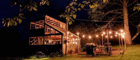 Et toetagers træhus med store vinduer og en terrasse, omgivet af en græsplæne og et træ.
