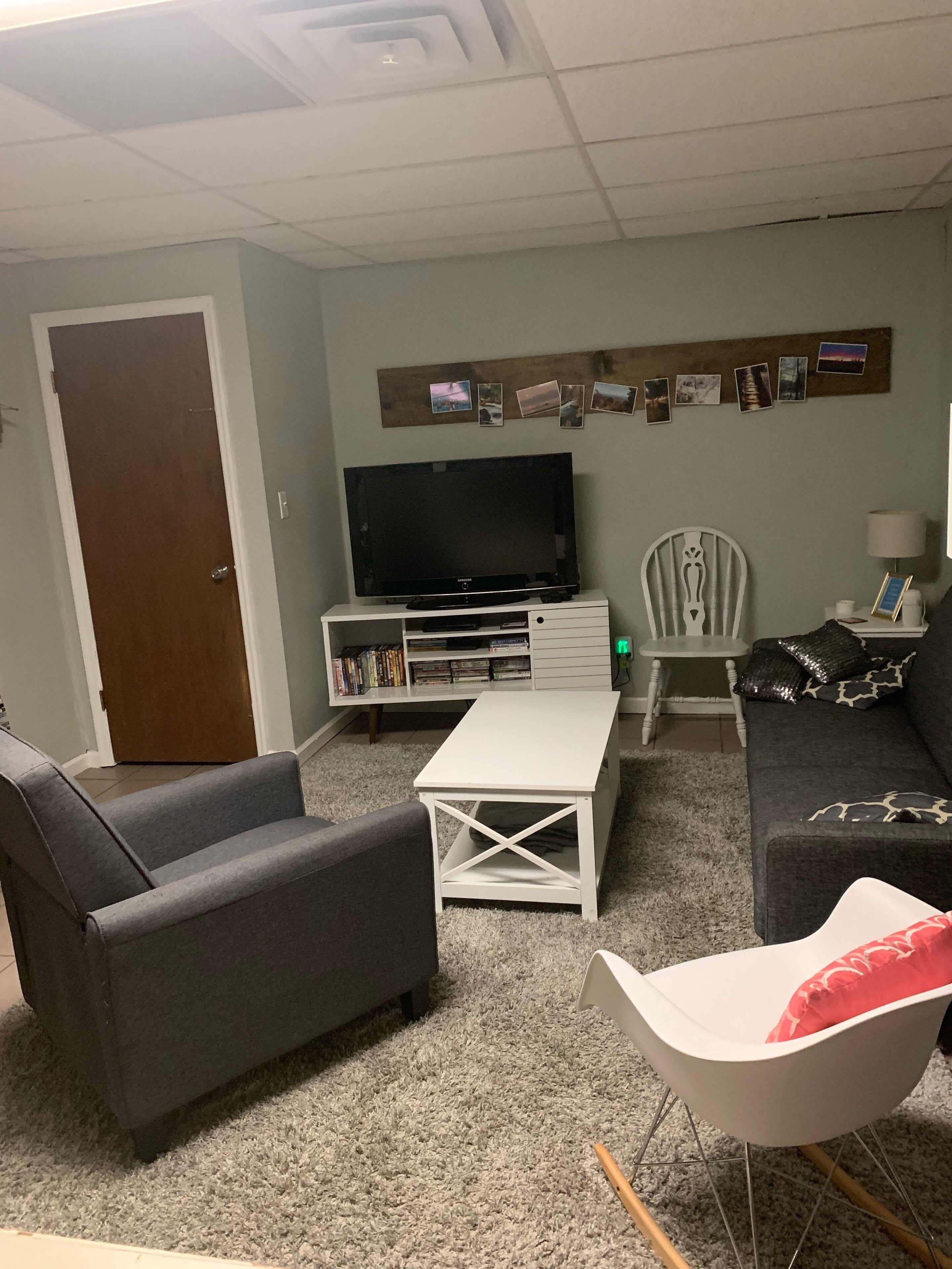 Una sala de estar con un televisor de pantalla plana, una estantería, una mesa de centro blanca, un sofá gris, un sillón blanco y un sofá gris.