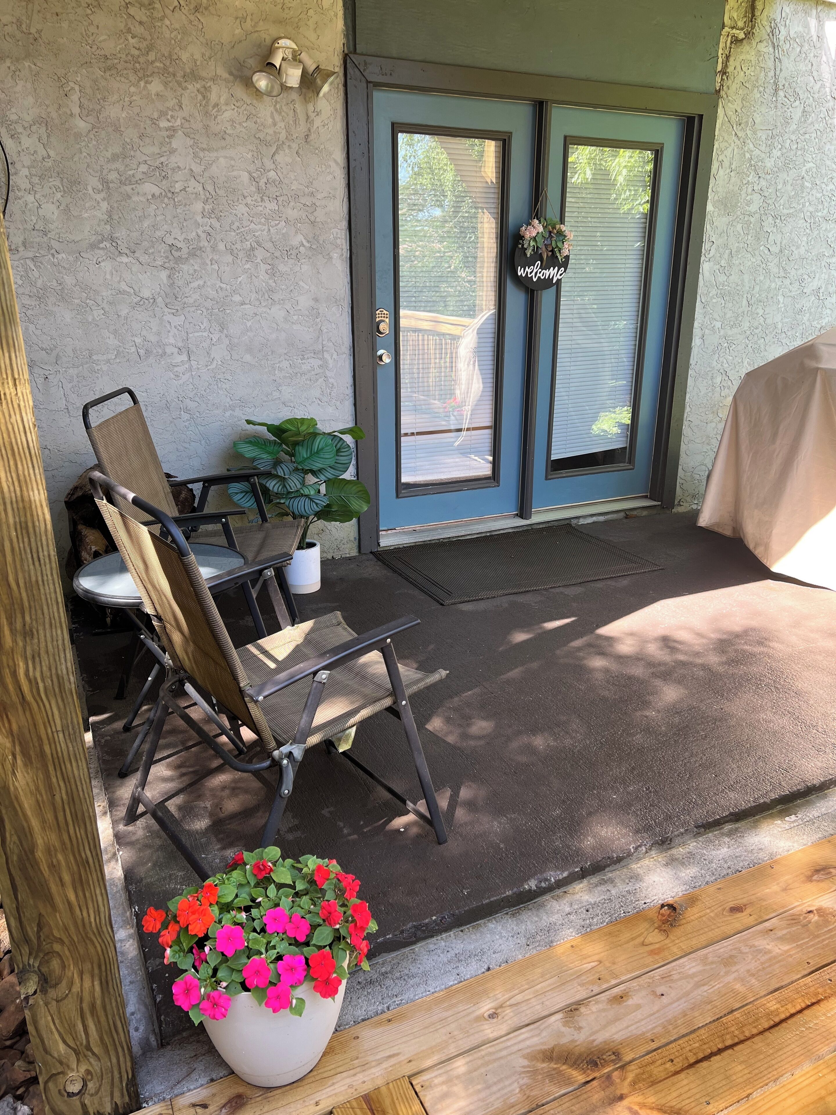 A covered patio area with a table and chairs, a potted plant, and a welcome sign on the door.