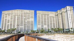 Et højt hotel med flere balkoner, en træpromenade, der fører til indgangen, og en klar blå himmel.