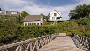 A wooden boardwalk leading to a white house with a green roof, surrounded by greenery and a clear sky. - Timeless Days, Oceanfront Fisherman’s Cottage, Deep Water Dock & Boathouse (Port Clyde)